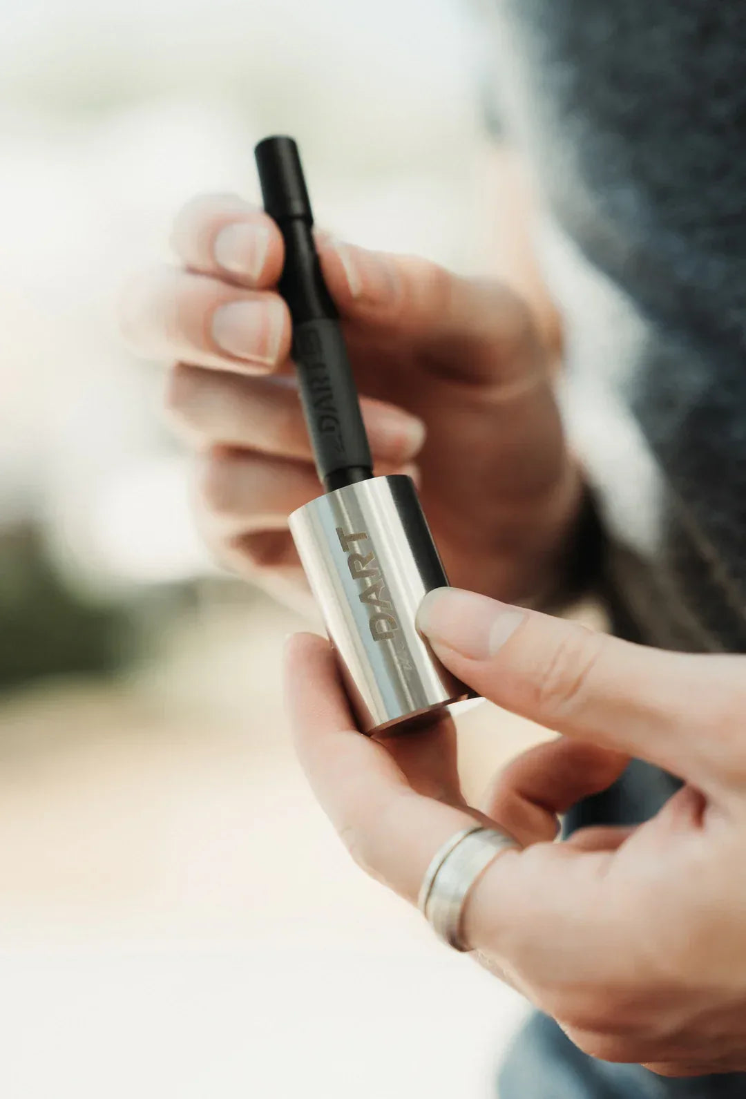 Hände halten einen silbernen „The Dart“-Behälter und führen eine schlanke, schwarze One-Hitter-Pfeife in die Öffnung ein.