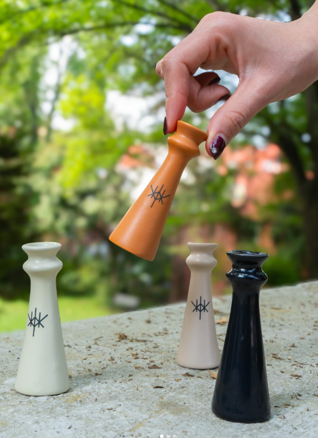 Eine Hand hält ein orangefarbenes, konisches Keramikgefäß über drei weitere in Weiß, Beige und Schwarz vor einem unscharfen grünen Hintergrund im Freien.