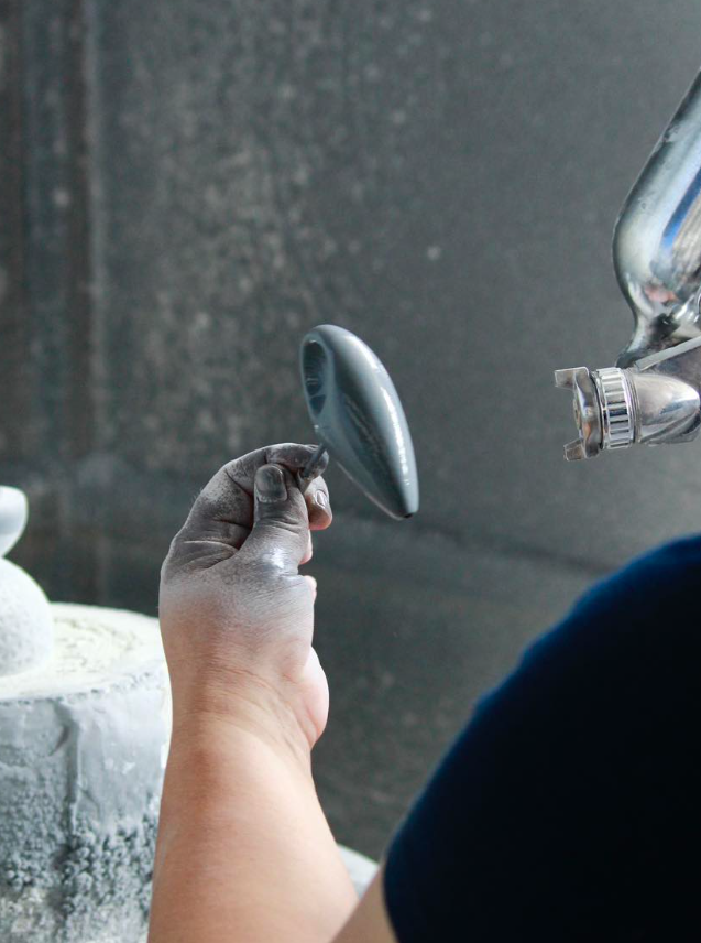 Die grau bestäubte Hand einer Person hält ein kleines, tropfenförmiges Keramikstück, während eine Spritzpistole zur Glasur verwendet wird.