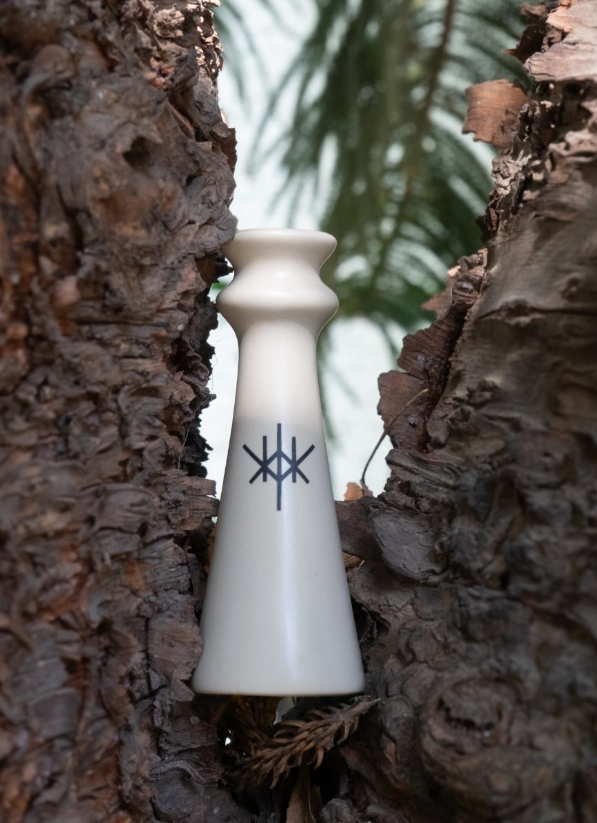 Ein cremefarbenes, konisches Keramikgefäß mit einem schwarzen geometrischen Symbol, eingebettet in die raue, dunkle Rinde eines Baumes.