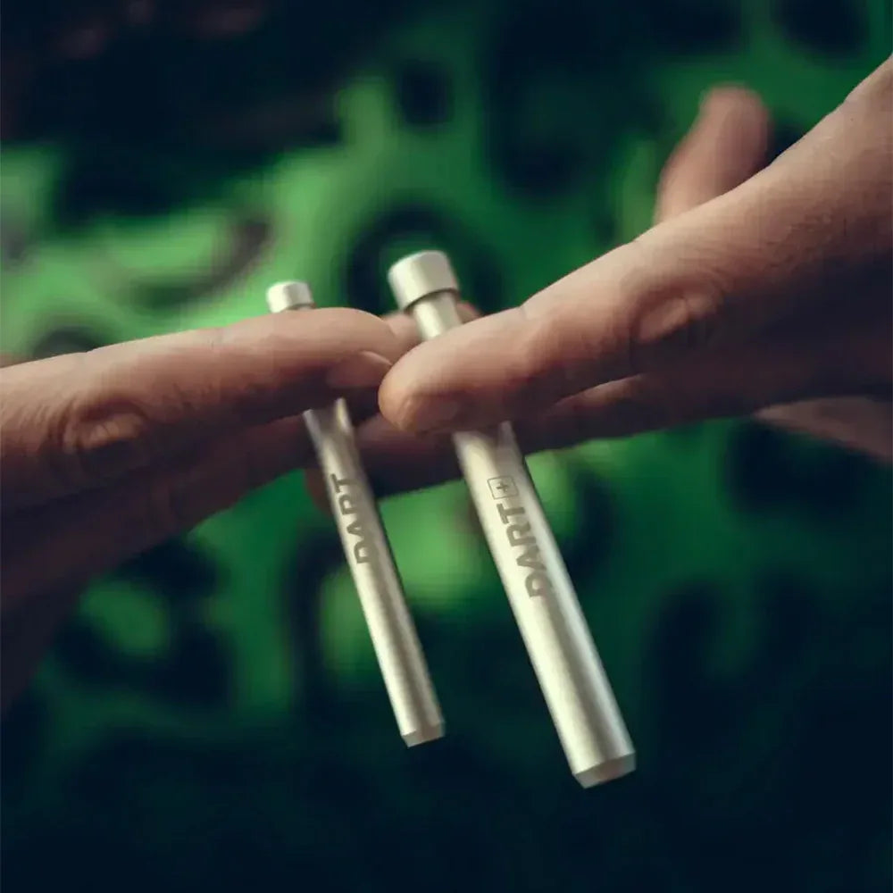 Zwei Hände halten eine silberne "The Dart" und eine größere "The Dart Plus" Pfeife nebeneinander vor einem verschwommenen grünen Hintergrund.