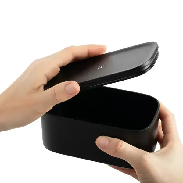 Zwei Hände, die den Deckel einer rechteckigen, mattschwarzen PAX-Aufbewahrungsdose öffnen, was deren kompakte Größe und glatte Oberfläche verdeutlicht.