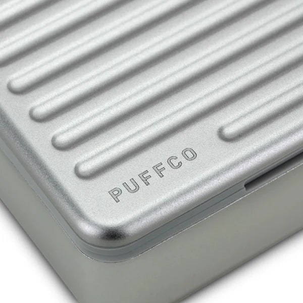 Eine Nahaufnahme des silbernen Puffco Travel Case, die das geprägte „PUFFCO“-Logo und die strukturierte, geriffelte Metalloberfläche zeigt.